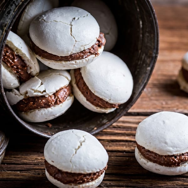 White meringue cookies with chocolate filling spilling from a dark metal pot onto a rustic wooden surface.