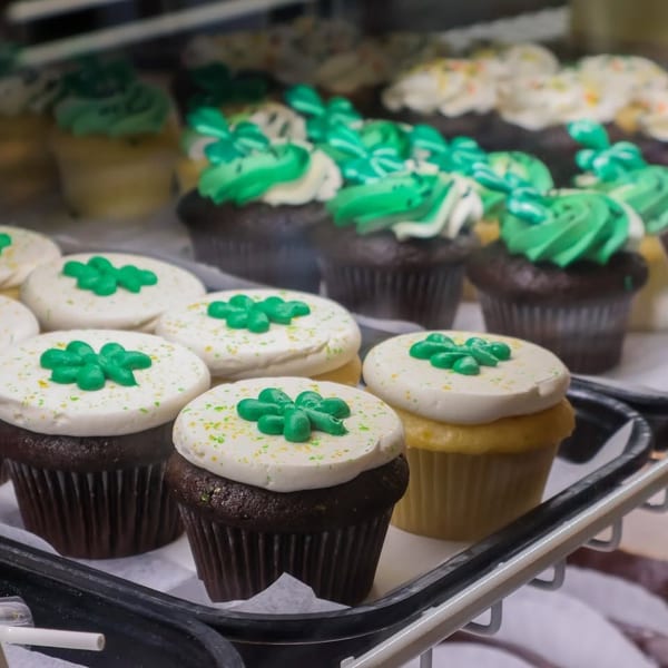 Cupcakes with white and green frosting, some topped with green shamrocks, displayed in a bakery case.