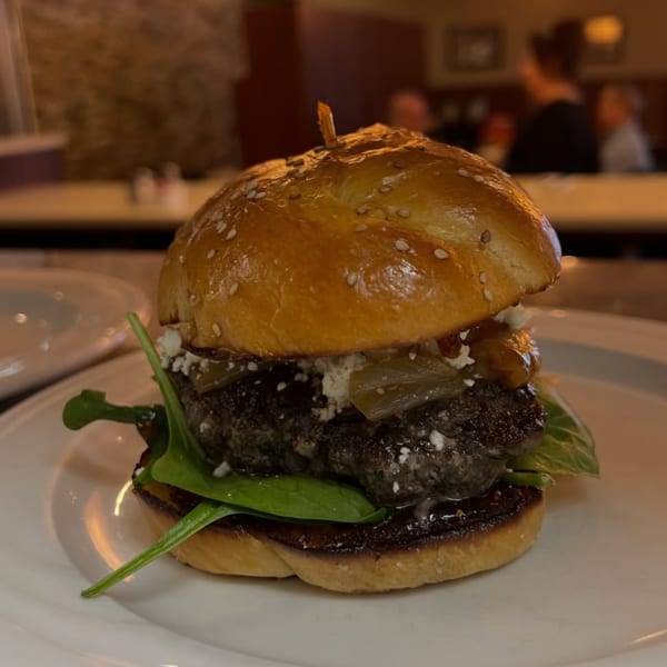 Gourmet beef burger with sesame brioche bun, greens, onions, and sauce, plated in a warmly lit restaurant.
