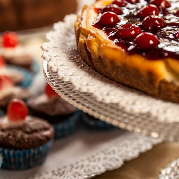 Cherry cheesecake on a glass stand next to chocolate cupcakes with cherries.