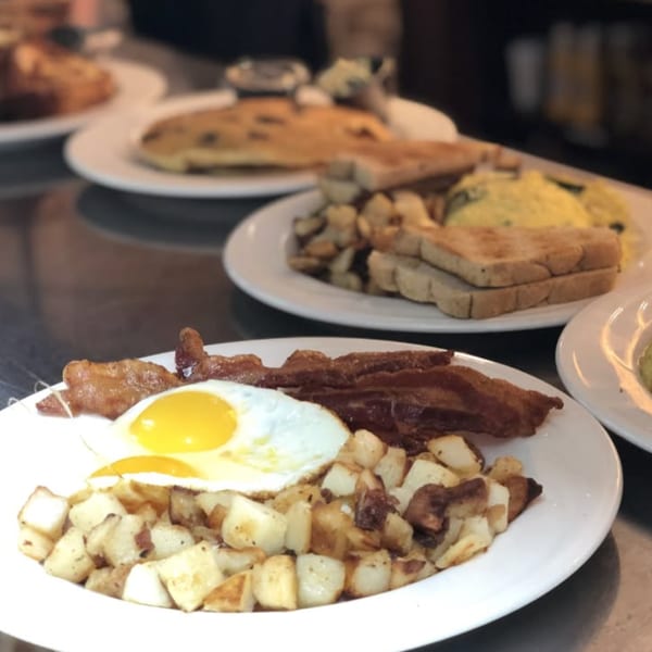 Breakfast plates featuring fried eggs, bacon, roasted potatoes, pancakes, sandwiches, and pastries on a diner counter.